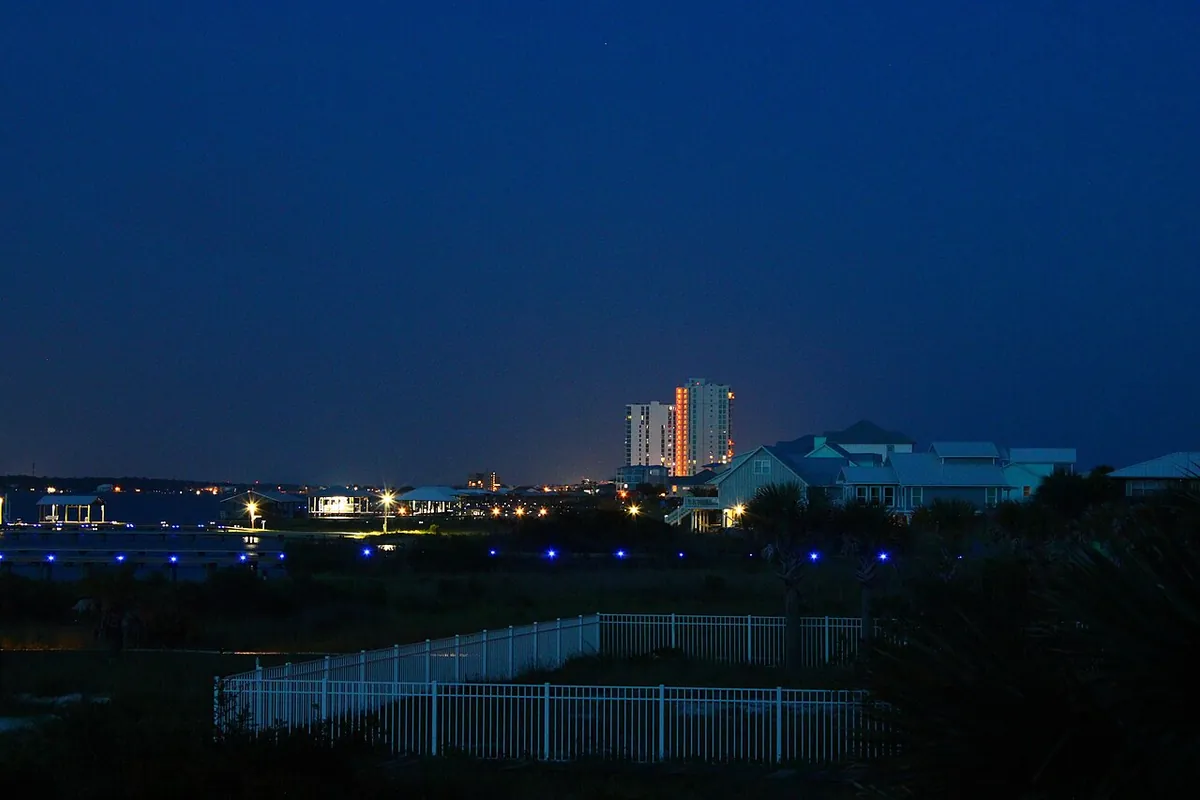 A lit beachfront condominium tower behind low-rise coastal houses at dusk in Gulf Shores AL